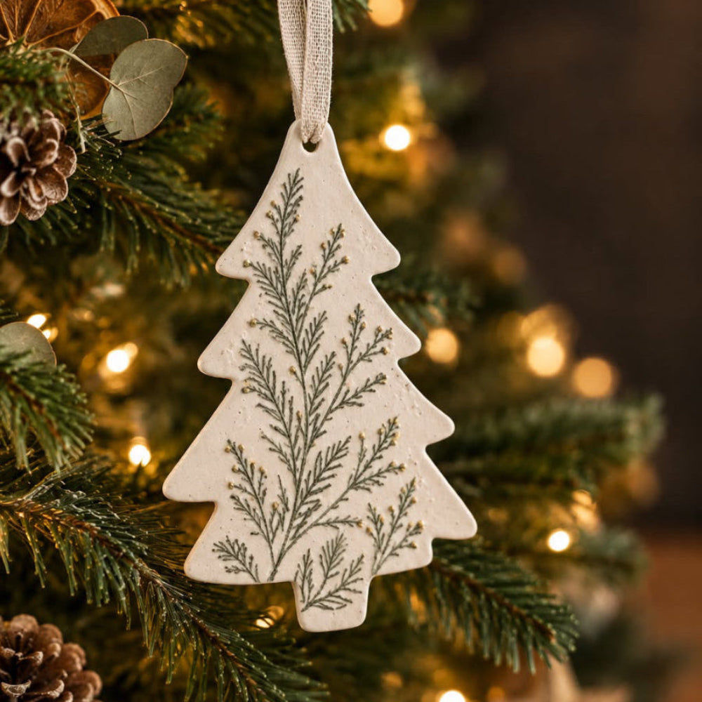 A Christmas tree ornament shaped like a pine tree, featuring a botanical branch pattern in olive green on an off-white background, hanging from a decorated Christmas tree with warm lights.